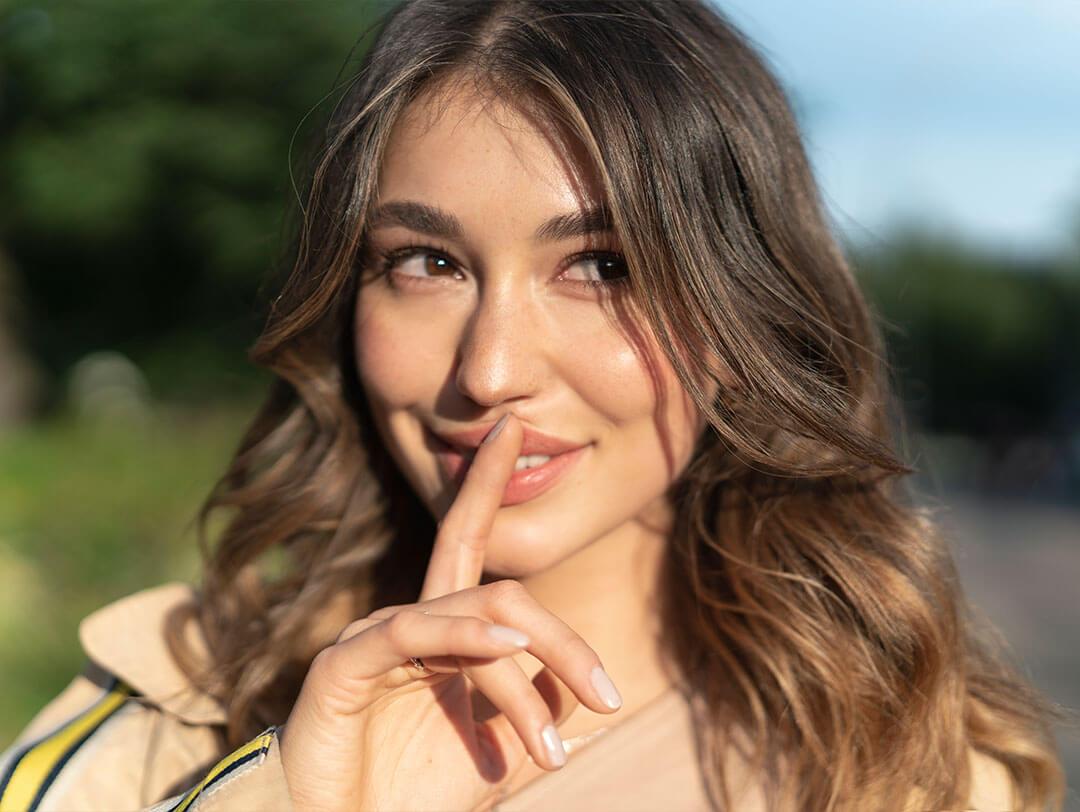 A close-up image capturing a smiling woman with her finger resting on the top of her lips